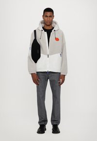 Felpa con fodera in pile, con zip, di colore grigio e bianco, con un'applicazione arancione, abbinata a jeans grigi, scarpe nere e una borsa a tracolla nera.