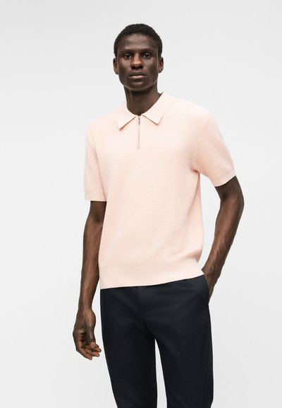 Άνδρας μοντέλο φοράει ροζ ανοιχτού χρώματος κοντομάνικο πόλο με υφασμάτινη υφή, γιακά με φερμουάρ και μαύρο παντελόνι μπροστά σε απλό ανοιχτό φόντο.
