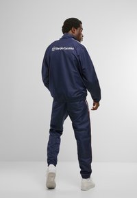 Námornícky športový overal s bielymi a červenými pruhmi, elastickým pásom a s yadačkou na zips. Na zadnej strane sa nachádza logo Sergio Tacchini.
