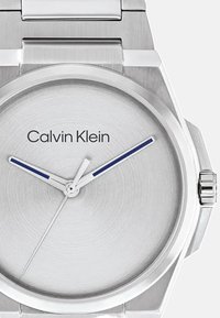 Srebrna ura iz nerjavnega jekla z brušeno površino, ki ima okroglo številčnico, modro uro in blagovno znamko "Calvin Klein" v črni barvi.