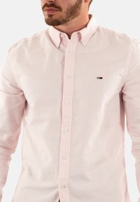 Chemise rose clair à manches longues avec de fines rayures blanches, col classique et petit logo brodé sur la poitrine gauche. Tissu en coton, coupe régulière.
