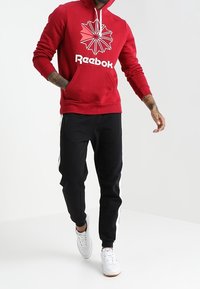 Sudadera roja con capucha y un logo blanco de Reebok junto a un diseño gráfico, combinada con joggers negros que tienen rayas laterales blancas y zapatillas blancas.