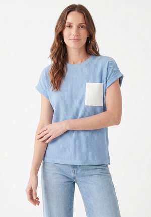 Kvinde med langt brunt hår iført en blå ribbet kortærmet top med hvid lomme og lyseblå jeans, stående foran en hvid baggrund.