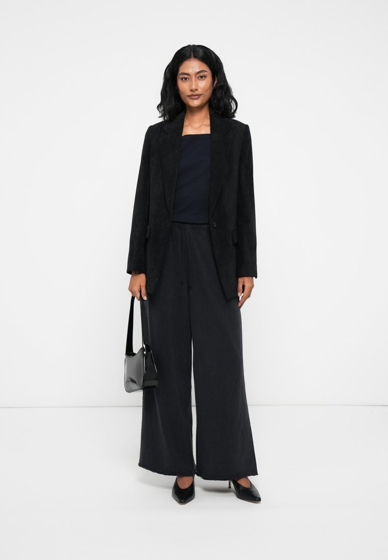 Blazer noir texturé sur un haut sombre, associé à un pantalon large noir. Les accessoires comprennent un sac à main noir et des chaussures à bouts pointus.