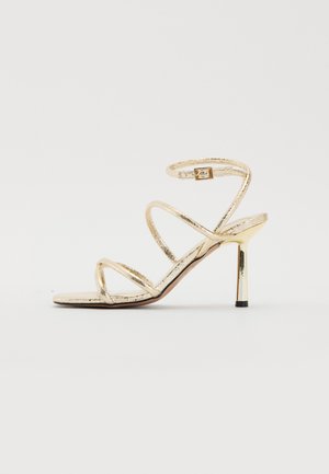 Zlaté páskové sandály se třemi diagonálními pásky, zapínáním na přezku a metalickým stiletto podpatkem. Hladký, lesklý povrch po celém modelu.