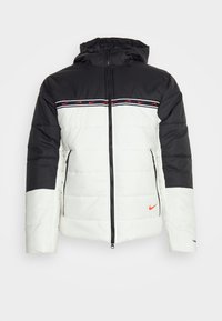 Chaqueta acolchada con bloques en blanco y negro, logo de Nike en naranja, cierre con cremallera en la parte delantera, capucha ajustable y detalles a rayas en la parte superior del pecho.