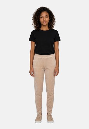 Donna con capelli ricci in piedi di fronte, indossando una maglietta nera, pantaloni beige e sneakers beige su uno sfondo semplice.