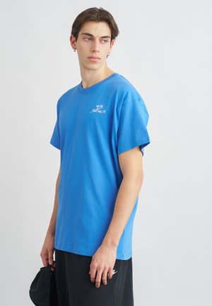 Blauwe katoenen t-shirt met korte mouwen, voorzien van een witte "Just Do It" logo. Draagt met zwarte shorts, weergegeven tegen een effen achtergrond.