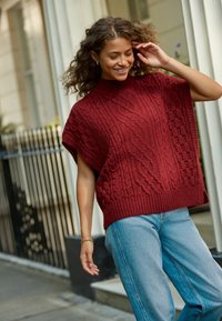 Pull en maille bordeaux à col montant, avec des motifs tressés et des manches courtes, assorti à un jean bleu clair.