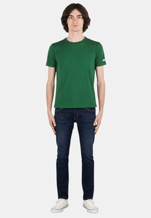 Giovane uomo in piedi di fronte, indossa una maglietta verde a maniche corte, jeans blu scuro e sneakers bianche su uno sfondo semplice.