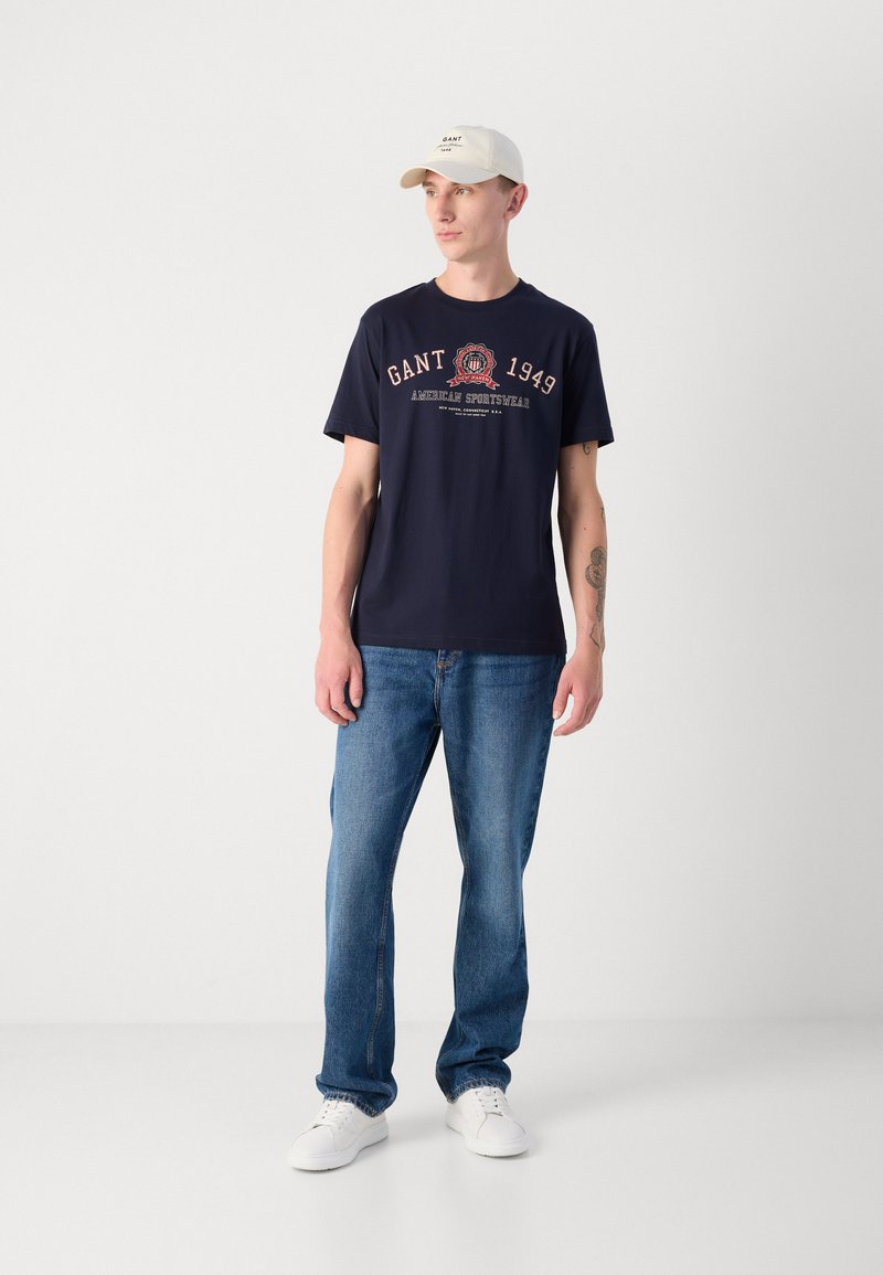 Granatowa bawełniana koszulka z nadrukiem "GANT 1949", w zestawie z niebieskimi dżinsami i białymi sneakersami, uzupełniona białą czapką.