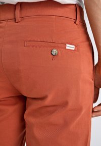 Rudawobrązowe spodnie z tylną kieszenią z zapięciem na guzik oraz metką. Materiał jest gładki z subtelną teksturą.