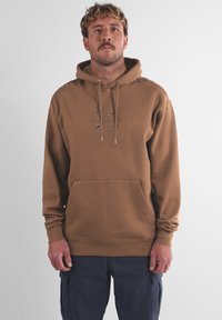 Sweat à capuche marron en tissu doux ; comporte une poche kangourou, un cordon de serrage et un logo embossé sur la poitrine.