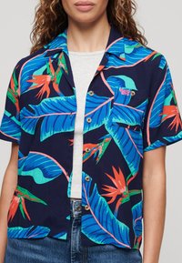Korte mouw shirt van stof met een marineblauwe achtergrond en levendige tropische bladeren en bloemen in teal, rood en roze. Met een knoopsluiting aan de voorkant.