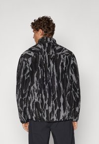 Veste en peau de mouton noire et grise avec un motif texturé et rayé. Elle possède un col montant et une coupe décontractée. La vue arrière montre des détails de couture.