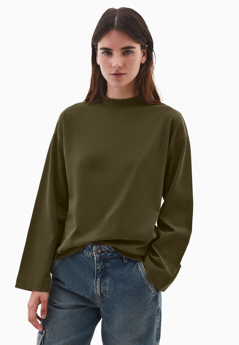 Oliwkowa bluzka z długim rękawem i wysokim kołnierzem, wykonana z miękkiego materiału. Charakteryzuje się luźnym krojem i opadającymi ramionami, zestawiona z niebieskimi dżinsami.