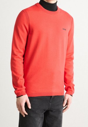 Mężczyzna ubrany w jaskrawoczerwony sweter z długim rękawem z logo "BOSS" na piersi, nałożony na czarny golf, połączony z ciemnymi jeansami.