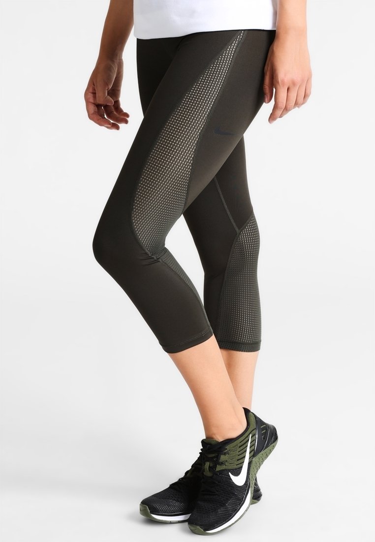 Personne portant des leggings noirs et vert olive courts avec des panneaux en mesh et des chaussures de course Nike noires et vertes, debout sur un fond blanc.