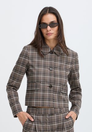 Femme portant des lunettes de soleil noires et une veste de costume à carreaux marron avec pantalon assorti, debout les mains dans les poches devant un fond uni.