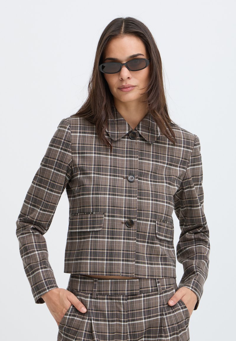 Femme portant des lunettes de soleil noires et une veste de costume à carreaux marron avec pantalon assorti, debout les mains dans les poches devant un fond uni.