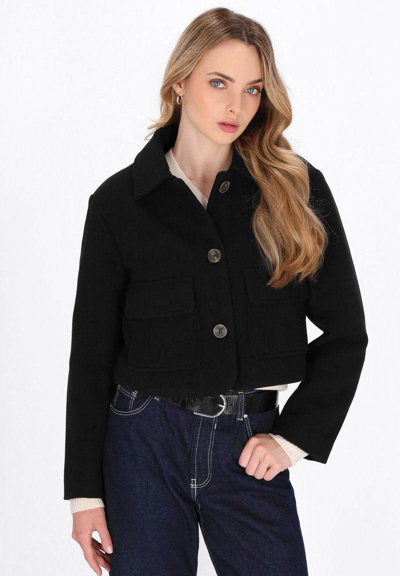 Kvinde med langt blond hår iført en sort knapjakke med store lommer, hvid top og mørkeblå højtaljede jeans med bælte.