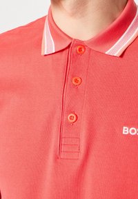 Gros plan sur une personne portant un polo rouge corail avec trois boutons fermés et un détail de col à rayures blanches.