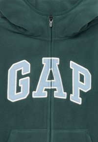 Sweat à capuche vert en polaire avec fermeture éclair sur le devant, ornée du mot "GAP" brodé en bleu clair et blanc. Comprend des poches kangourou.
