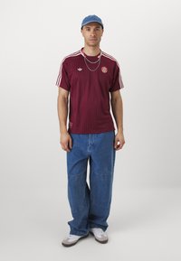 Maglia Adidas a righe bordeaux con maniche corte, logo e distintivo sul petto. Indossata con jeans larghi blu chiaro e sneaker bianche.