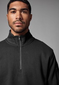 Pullover nero con zip alta, realizzato in tessuto testurizzato. Presenta una zip di lunghezza quarti e una tasca laterale con zip.