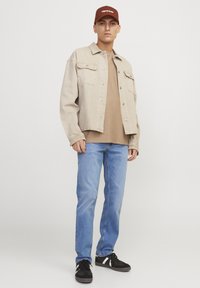 Chaqueta de mezclilla beige, camiseta marrón, jeans azul claro y zapatillas negras con detalles blancos. La gorra es color burdeos con texto blanco.