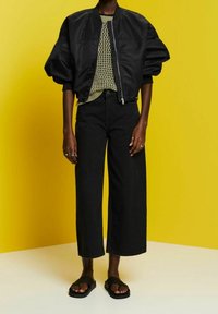 Personne portant un pantalon large noir court, une veste bomber noire oversize, un haut en maille verte, des sandales noires, debout sur un fond jaune.