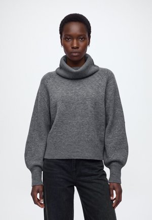 Vrouw met kort haar draagt een grijze coltrui en zwarte broek, staand tegen een effen witte achtergrond.