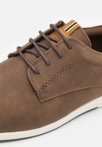 Zapato de cuero marrón con un acabado texturizado, que presenta cordones marrones, un acento a rayas amarillo y negro en contraste, y una suela blanca.