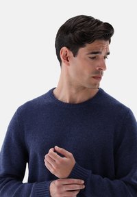 Maglione a girocollo blu navy realizzato in tessuto morbido con polsini e orlo a costine. Superficie liscia, design aderente, con le mani incrociate davanti.