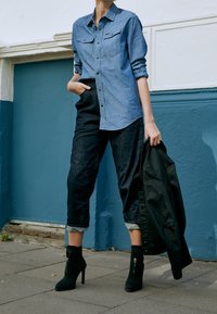 Person som bär en blå knappad skjorta och rullade mörka jeans, håller en svart jacka, står på en trottoar mot en blå och vit vägg.