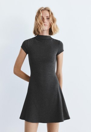 Szőke hajú nő, aki testhezálló, szénszürke, rövid ujjú, magas nyakú ruhát visel, egyszerű világos háttér előtt áll.