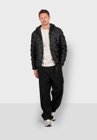 Mand iført sort quilted jakke, hvid skjorte, løse sorte bukser og hvide sneakers, står op ad en ensfarvet grå baggrund.