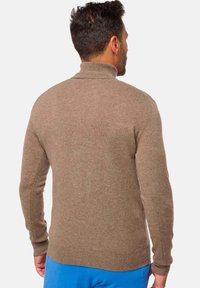 Mann, der mit dem Rücken zur Kamera steht, trägt einen braunen Rollkragenpullover und blaue Hosen vor einem schlichten hellen Hintergrund.