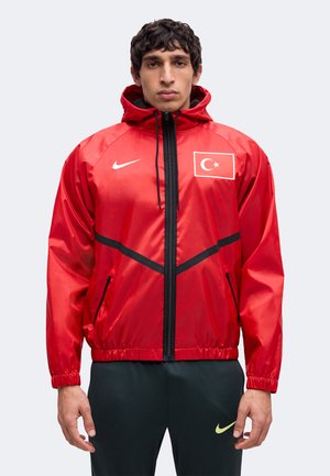 Άνδρας που φοράει κόκκινο μπουφάν Nike με την τουρκική σημαία και μαύρο φερμουάρ, στέκεται μπροστά σε απλό φωτεινό φόντο.
