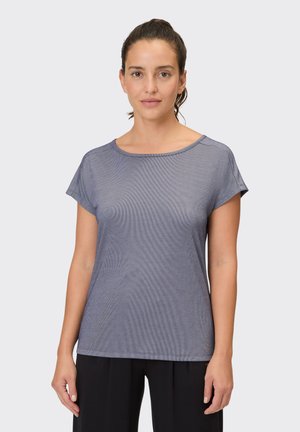 Woman with tied-back brown hair wearing a short-sleeve gray top and black pants, standing against a plain light background.