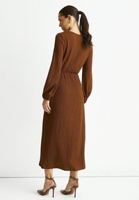 Rust-colored midi dress with long sleeves, textured fabric, and a cinched waist. Paired with transparent heeled sandals.