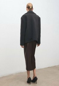 Abrigo de lana gris oscuro de gran tamaño con un gran cuello, dos botones en el dobladillo, combinado con una falda marrón hasta la rodilla y tacones de aguja negros.