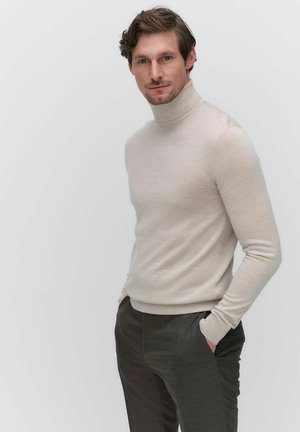 Jasno beżowy sweter z golfem wykonany z miękkiego materiału, dopasowany krój z długimi rękawami, noszony z ciemnoszarymi spodniami o eleganckim kroju, minimalistyczne tło.