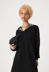 Črnaobleka z dolgimi rokavi in ovratnikom v leopardjem vzorcu, dopolnjena s teksturirano črno pleteno torbico. Preprost dizajn s V-izrezom.