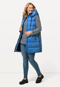 Blå vadderad väst med huva, dragkedjestängning och sidofickor, bärs över en svart-vitrandig långärmad topp och blåjeans.