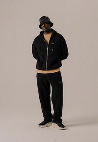 Homme debout portant un chapeau seau noir, un sweat à capuche noir zippé, une chemise beige, un pantalon noir et des baskets noires sur un fond beige.