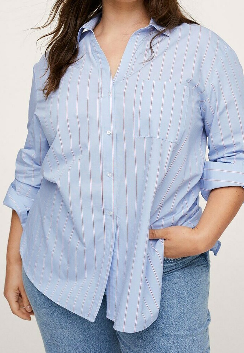Femme portant une chemise bleu clair à boutons avec de fines rayures rouges verticales et un jean bleu, une main dans la poche et les manches retroussées.