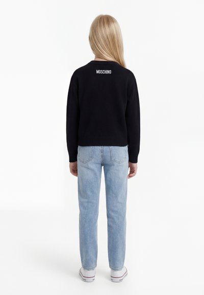 Barn med langt blond hår som har på seg en svart Moschino-genser, lyseblå jeans og hvite joggesko, vist bakfra.
