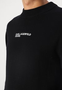Juodas megztinis iš mezgimo audinio su šonkačio apykakle. Ant krūtinės yra balta siuvinėta "KARL LAGERFELD JEANS" logo.