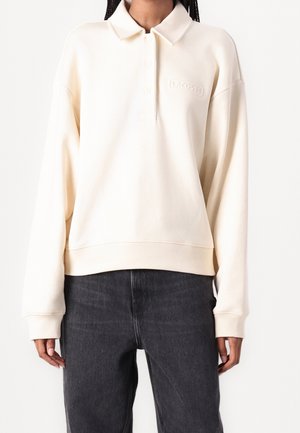 Persoon draagt een crèmekleurige polo sweat met lange mouwen en geborduurd "Lacoste"-logo en zwarte spijkerbroek tegen een effen witte achtergrond.